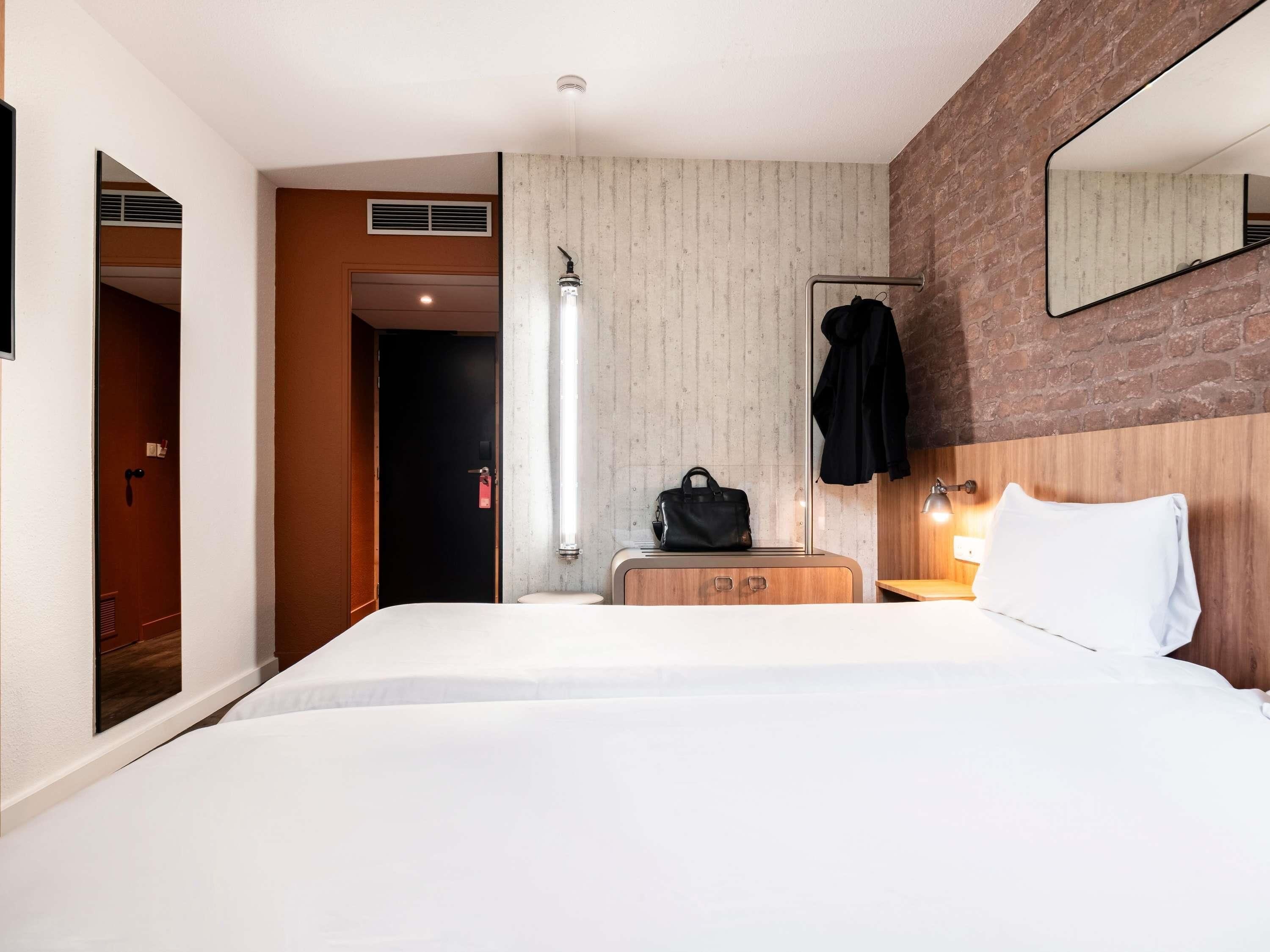 A hotel room with a bed, a wooden dresser, a mirror, a hanging coat, and a light fixture.