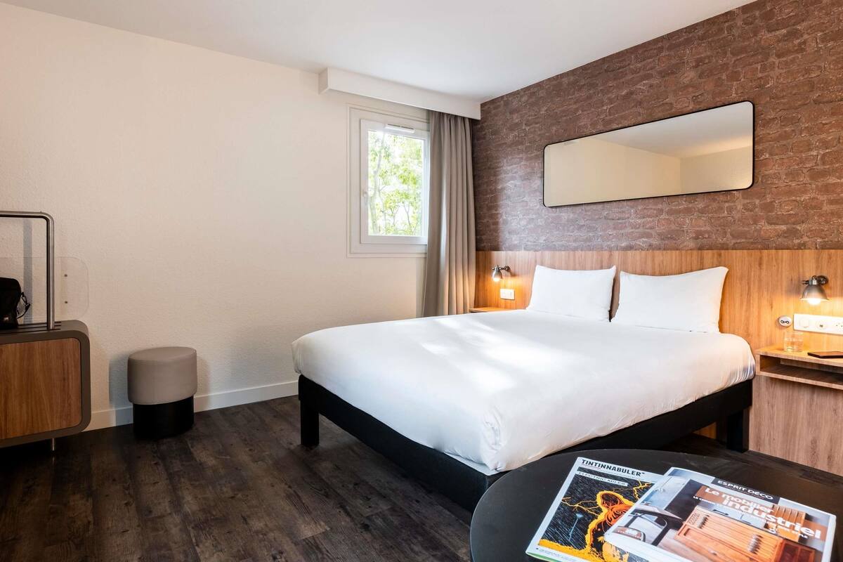 A hotel room with a large bed, a wooden headboard, a nightstand with a lamp, a small round table with a magazine, and a window with curtains.
