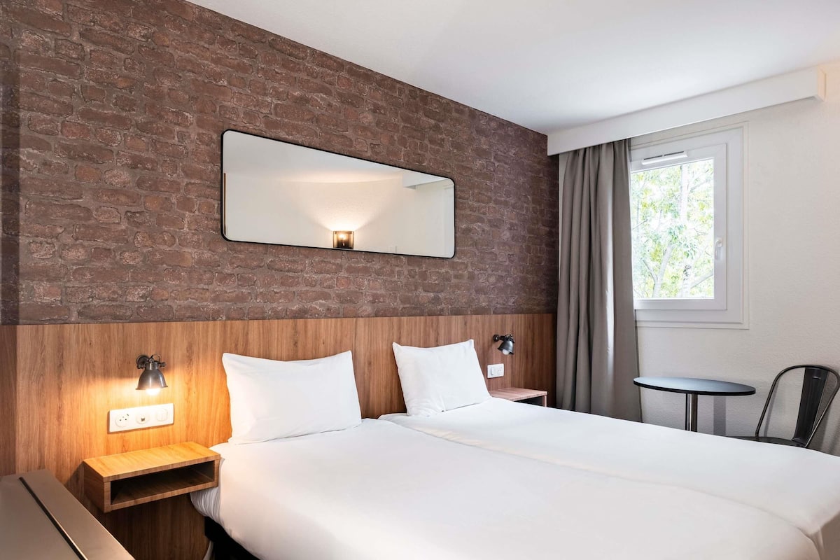 A hotel room with a brick wall, a large bed with white linens, a small table, and a chair.