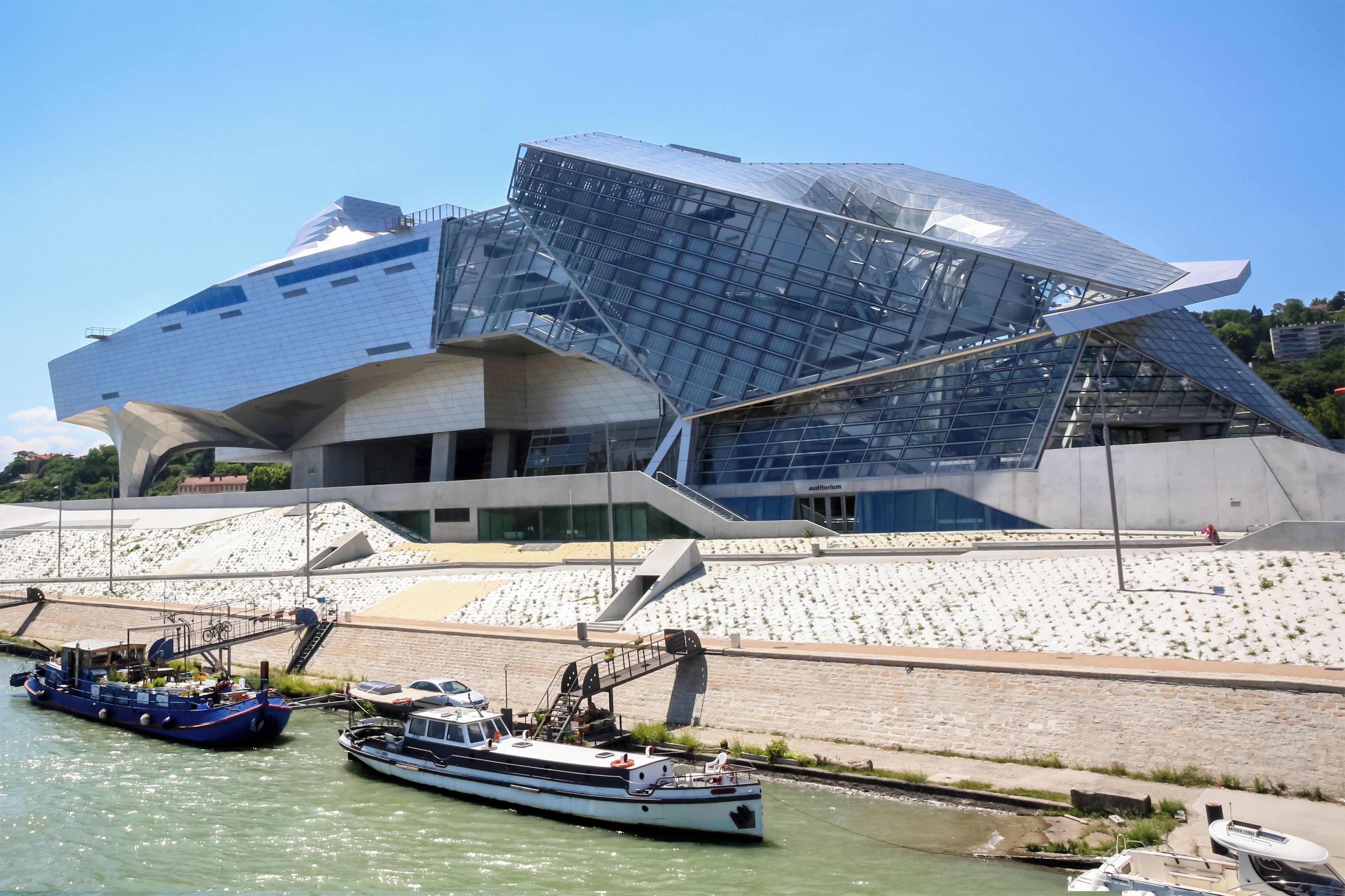A modern architectural building with a unique, angular design, situated by a river with boats docked nearby.