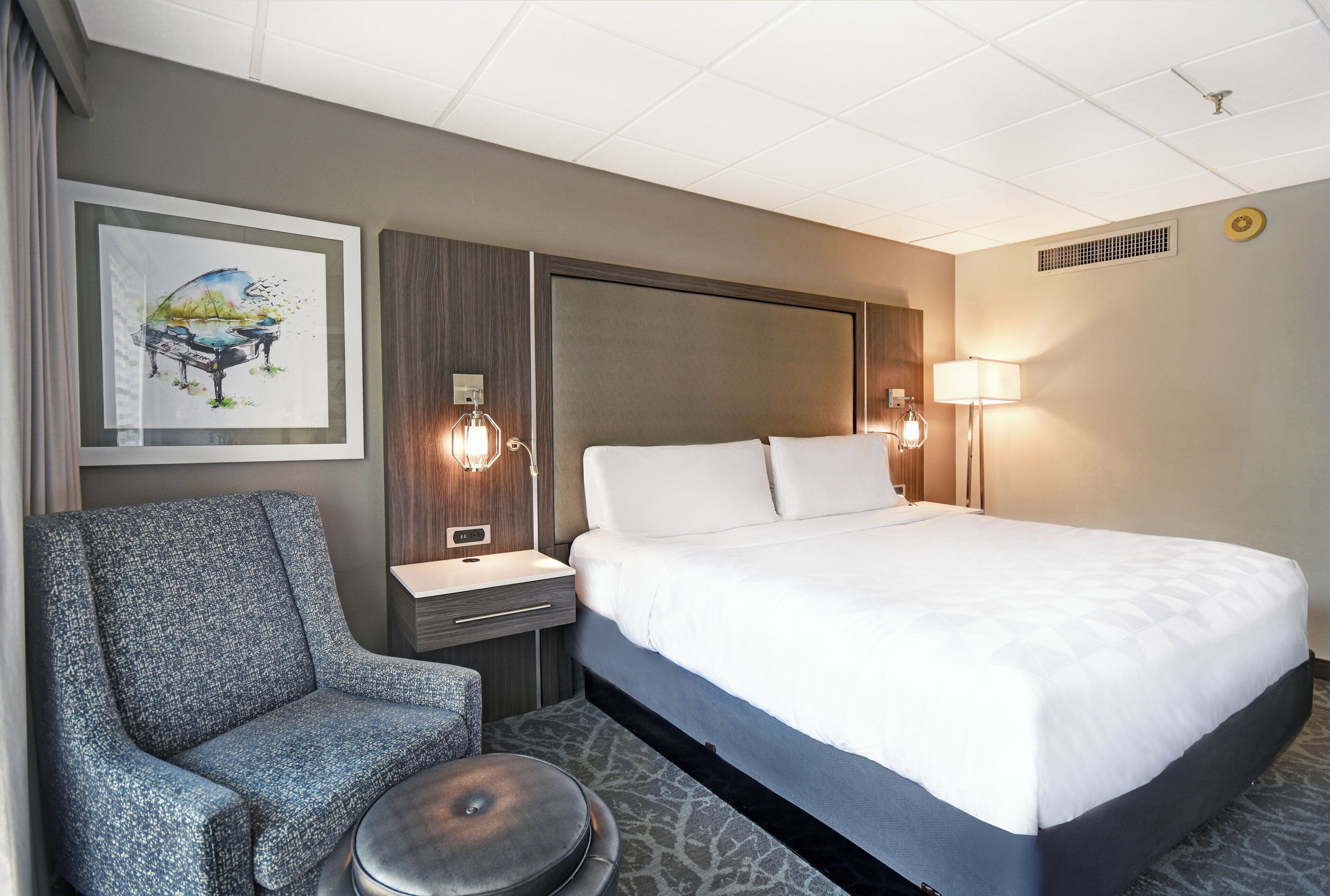 A hotel room with a bed, a chair, a nightstand, a lamp, and a painting on the wall.