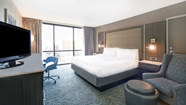 A hotel room with a large bed, a desk with a TV, a chair, a small ottoman, and a view of the city through large windows. - Holiday Inn Downtown Memphis by IHG (Memphis)