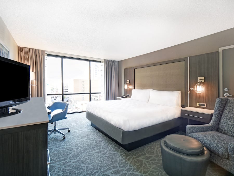 A hotel room with a large bed, a desk with a TV, a chair, a small ottoman, and a view of the city through large windows.