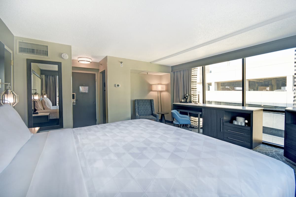 Habitación de hotel con una cama grande, un escritorio con silla, una mesita y vistas a la ciudad a través de amplios ventanales.