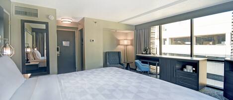 A hotel room with a large bed, a desk with a chair, a small table, and a view of the city through large windows.