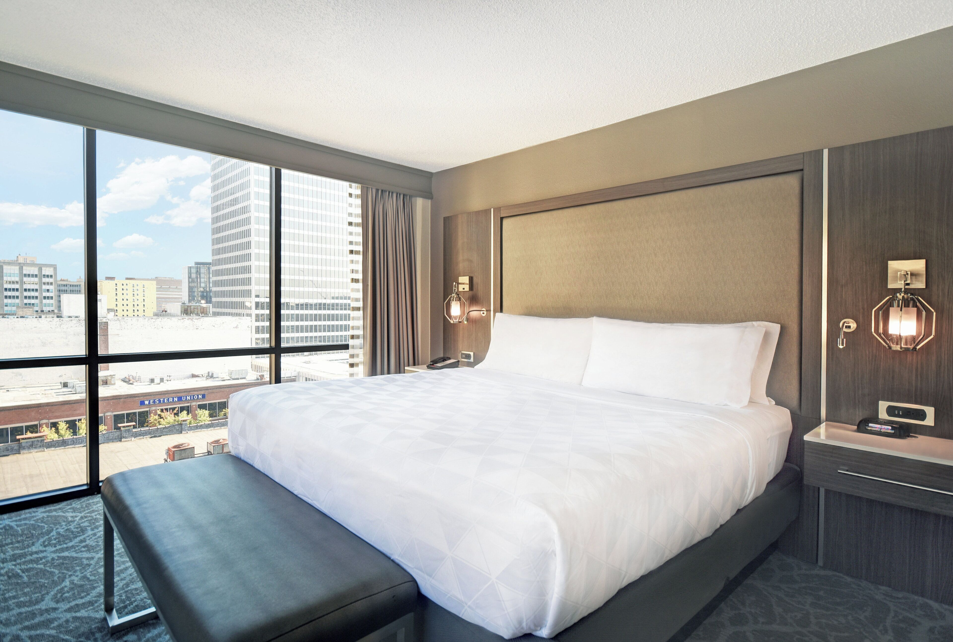 Een hotelkamer met een groot bed, een bankje, een nachtkastje, een wandlamp en uitzicht op de stadsgezicht door grote ramen.