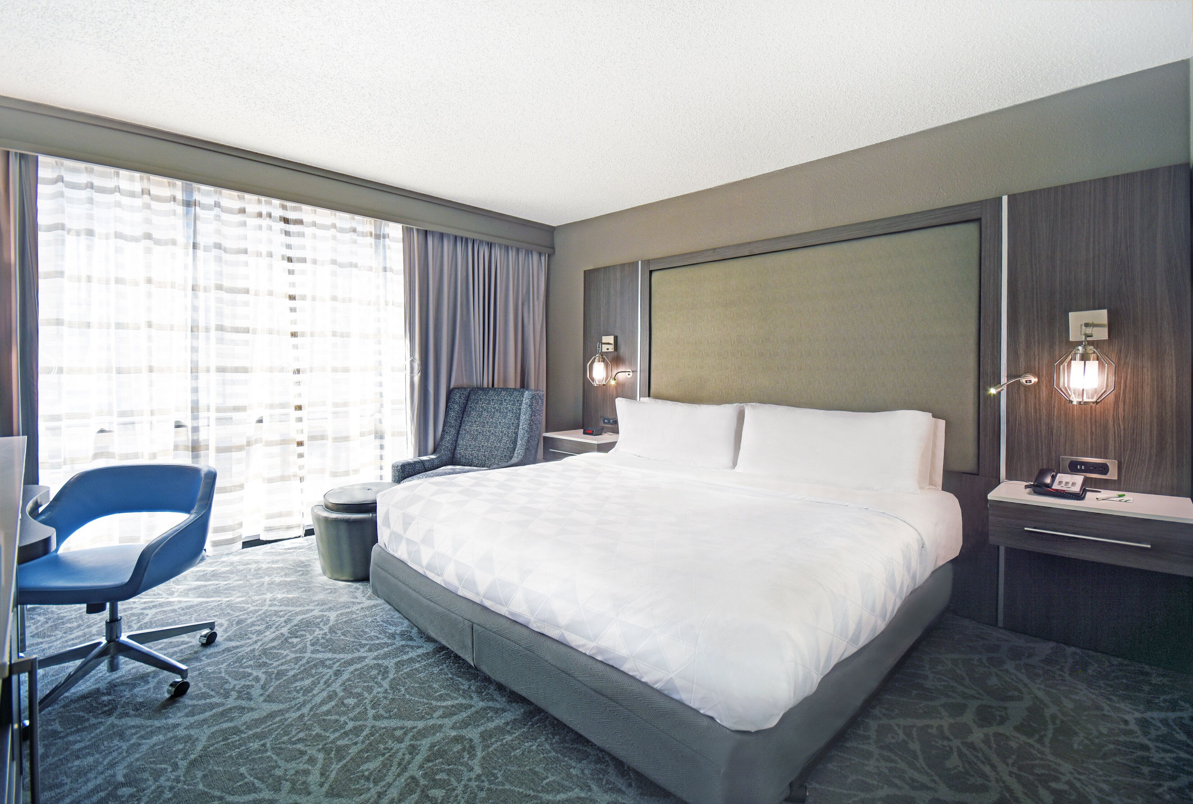 Quarto de hotel com uma cama grande, uma escrivaninha com cadeira, uma mesa de cabeceira com abajur e uma janela com cortinas.