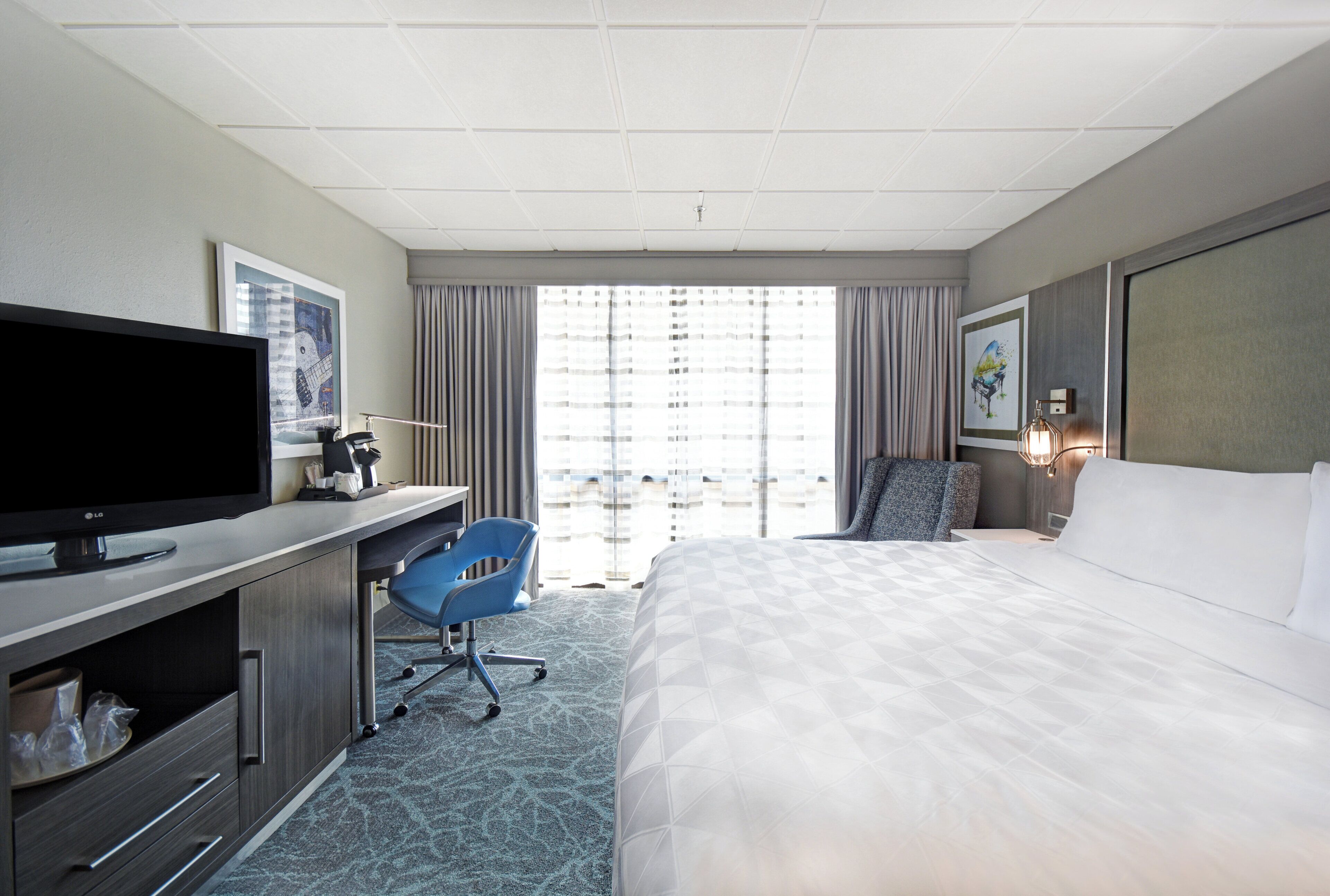 A hotel room with a bed, a desk with a flat-screen TV, a chair, a window with curtains, and artwork on the walls.