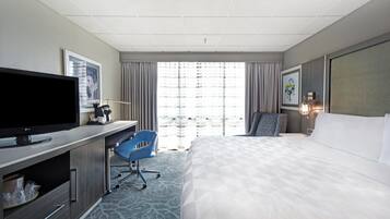Habitación de hotel con cama, escritorio con televisor de pantalla plana, una silla, ventana con cortinas y obras de arte en las paredes.