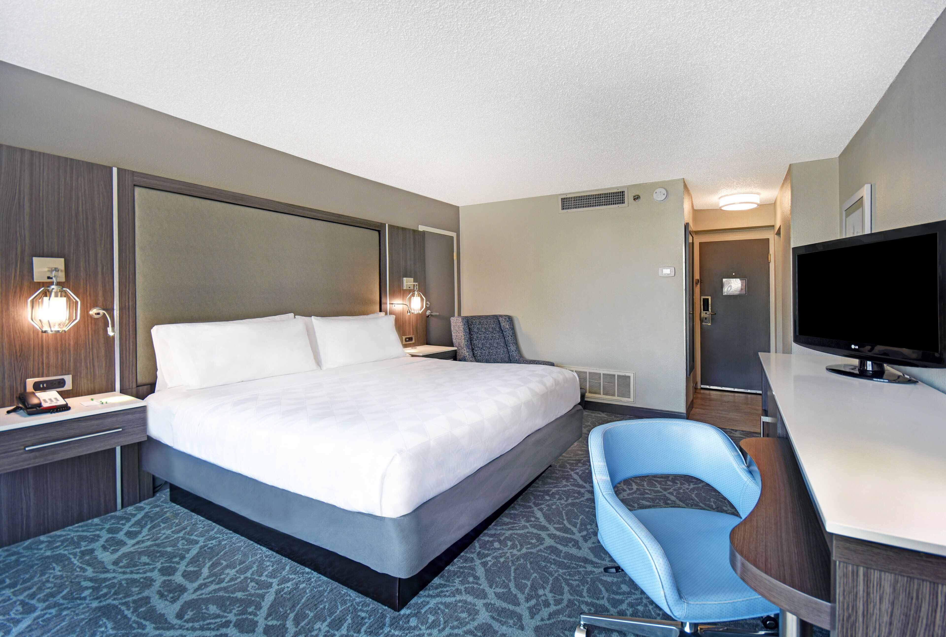 A hotel room with a large bed, a desk with a flat-screen TV, a blue chair, and a nightstand with a lamp.