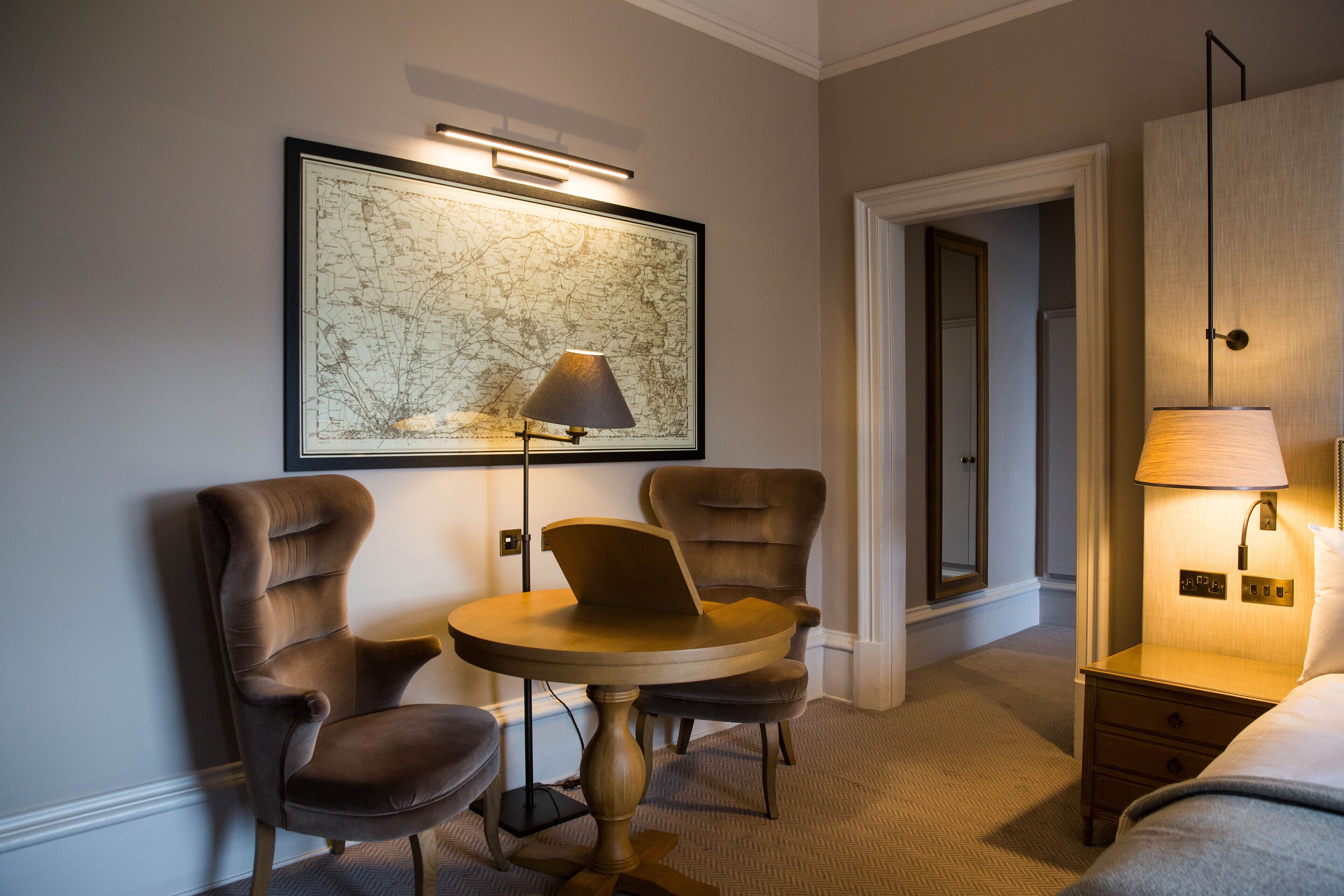 A hotel room with a round table, two armchairs, a large framed map on the wall, a bedside lamp, and a door leading to another room.