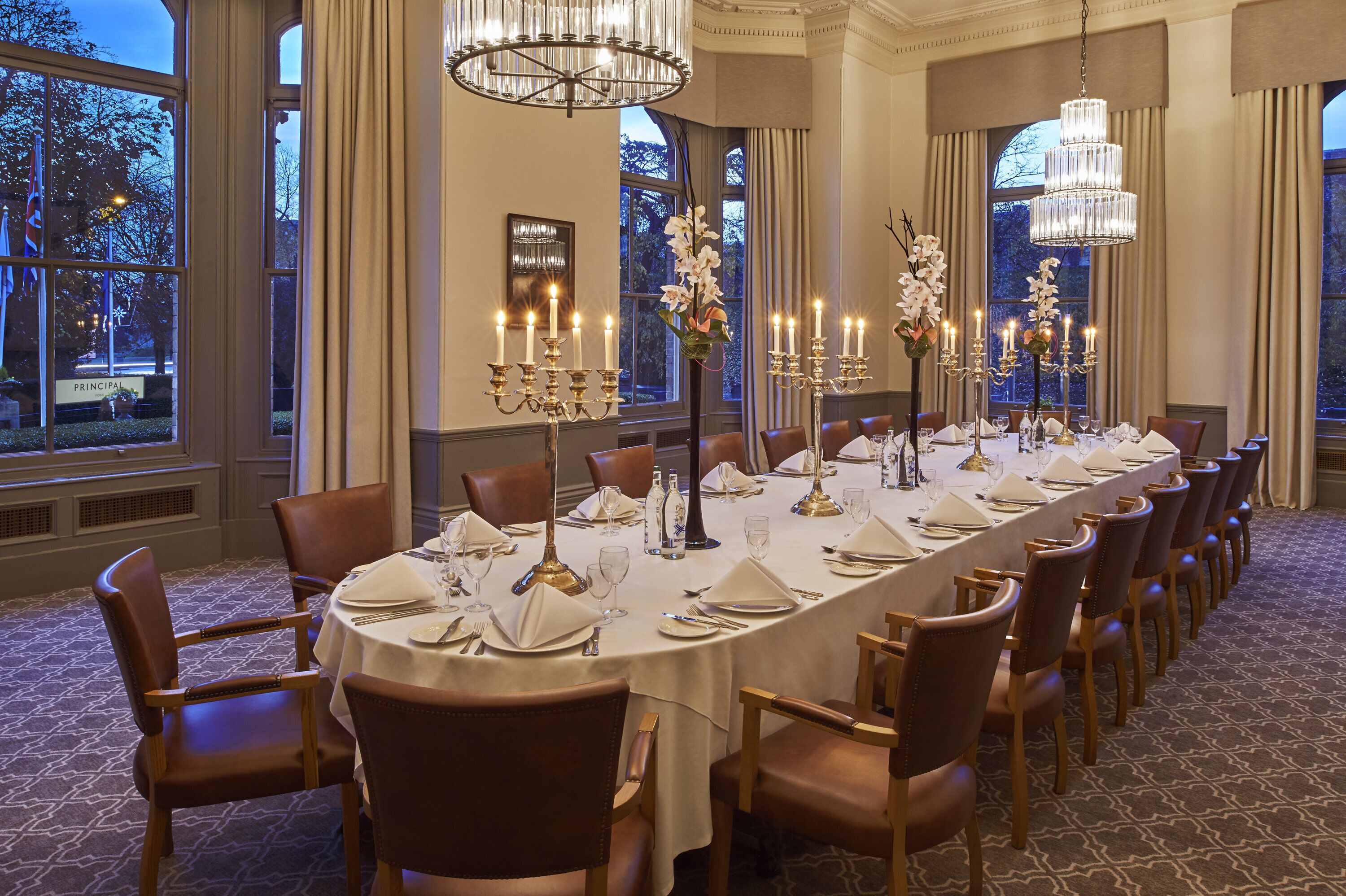 A formal dining room with a long table set for a meal, large windows, and elegant chandeliers.