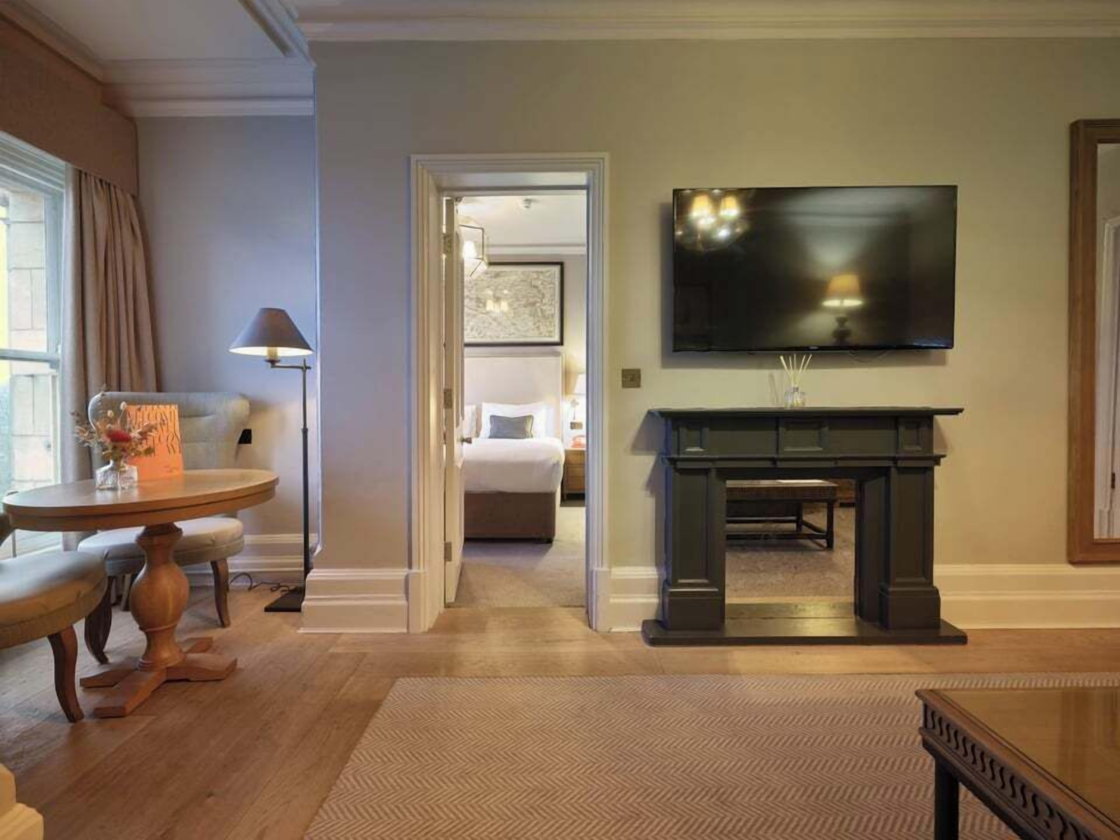 Ein Hotelzimmer mit großem Flachbildfernseher an der Wand, einem Holztischchen, einer Lampe und einem Bett mit Blick ins Zimmer.