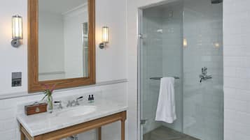 Un baño con lavamanos de mármol blanco, tocador de madera, un espejo grande y una ducha con cerramiento de vidrio.