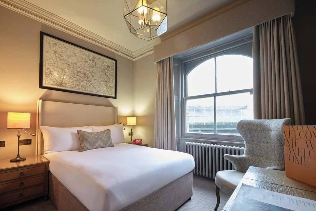 A hotel room with a large bed, a chair, a desk, a window with curtains, a radiator, and a framed artwork on the wall.