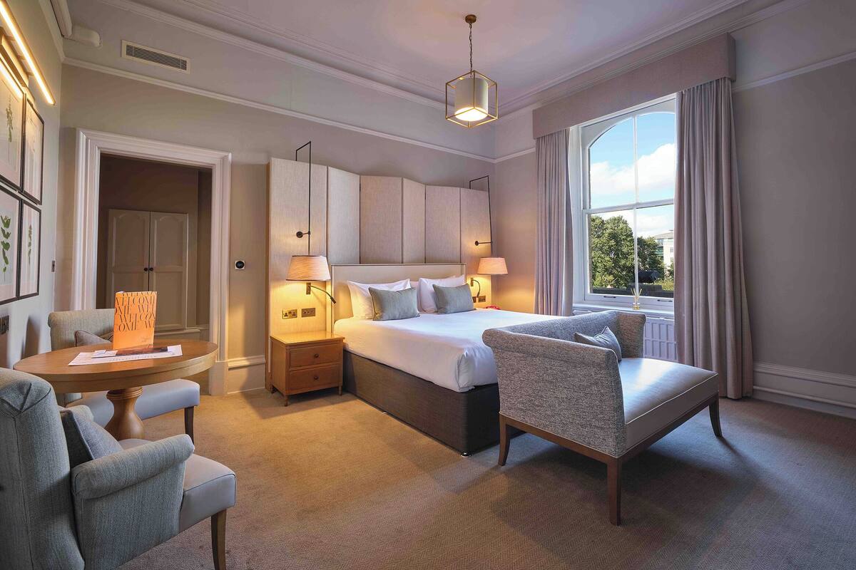 A hotel room with a large bed, two bedside tables with lamps, a sofa, a chair, and a large window with curtains.