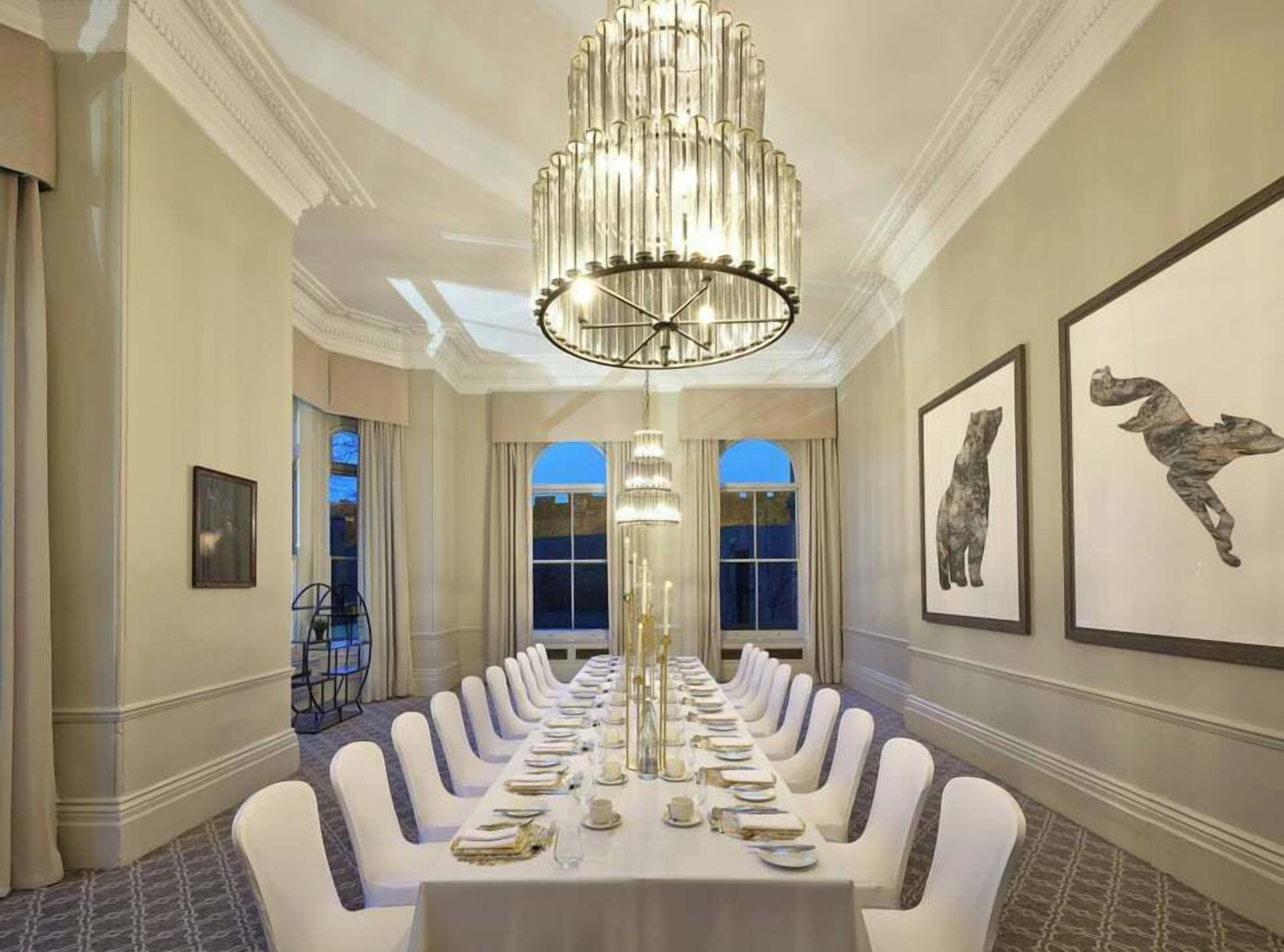 Une salle à manger formelle, avec une longue table dressée pour un repas, de magnifiques lustres et des œuvres d’art encadrées qui ornent les murs.