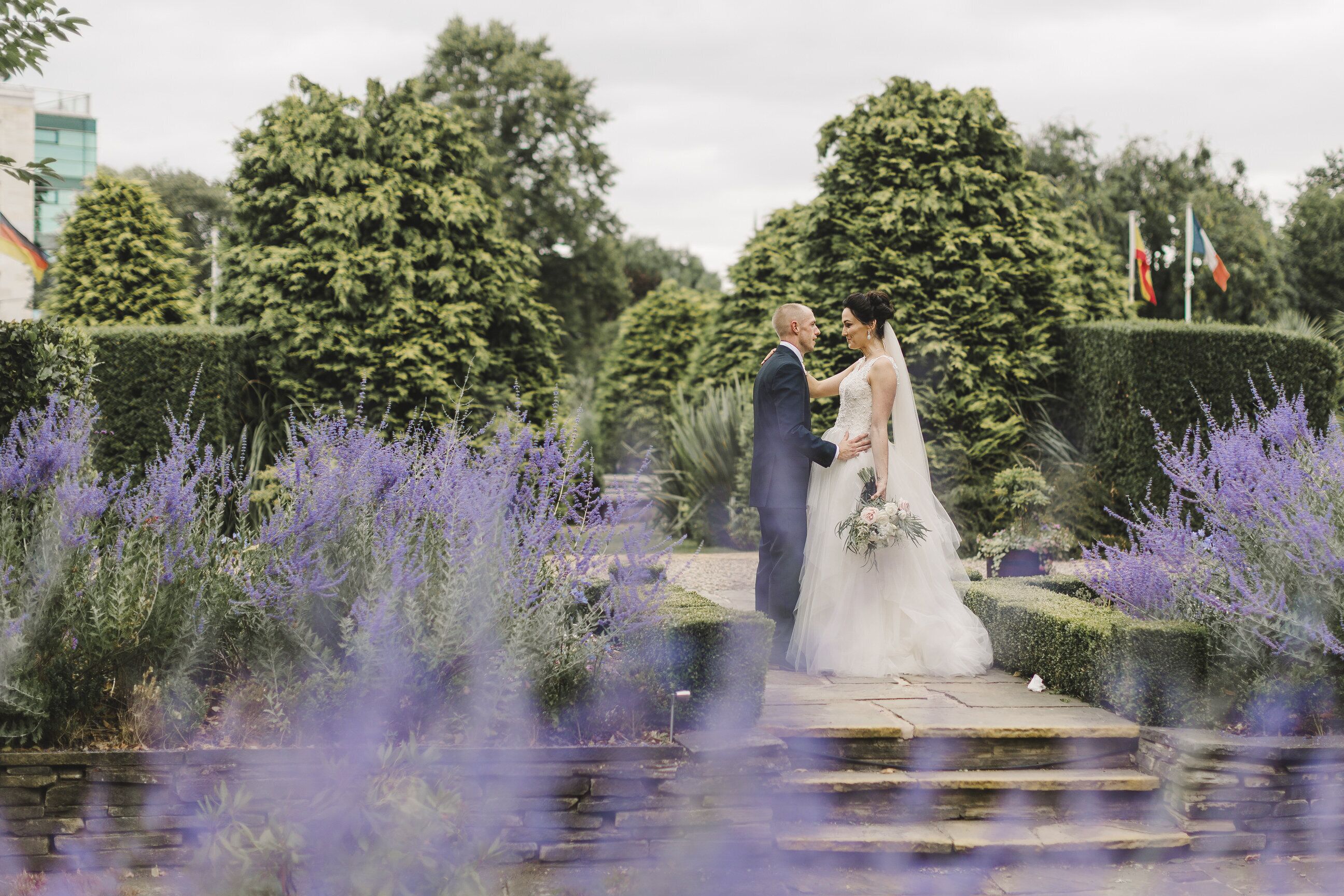 A couple standing on a stone pathway surrounded by lush greenery and purple flowers.