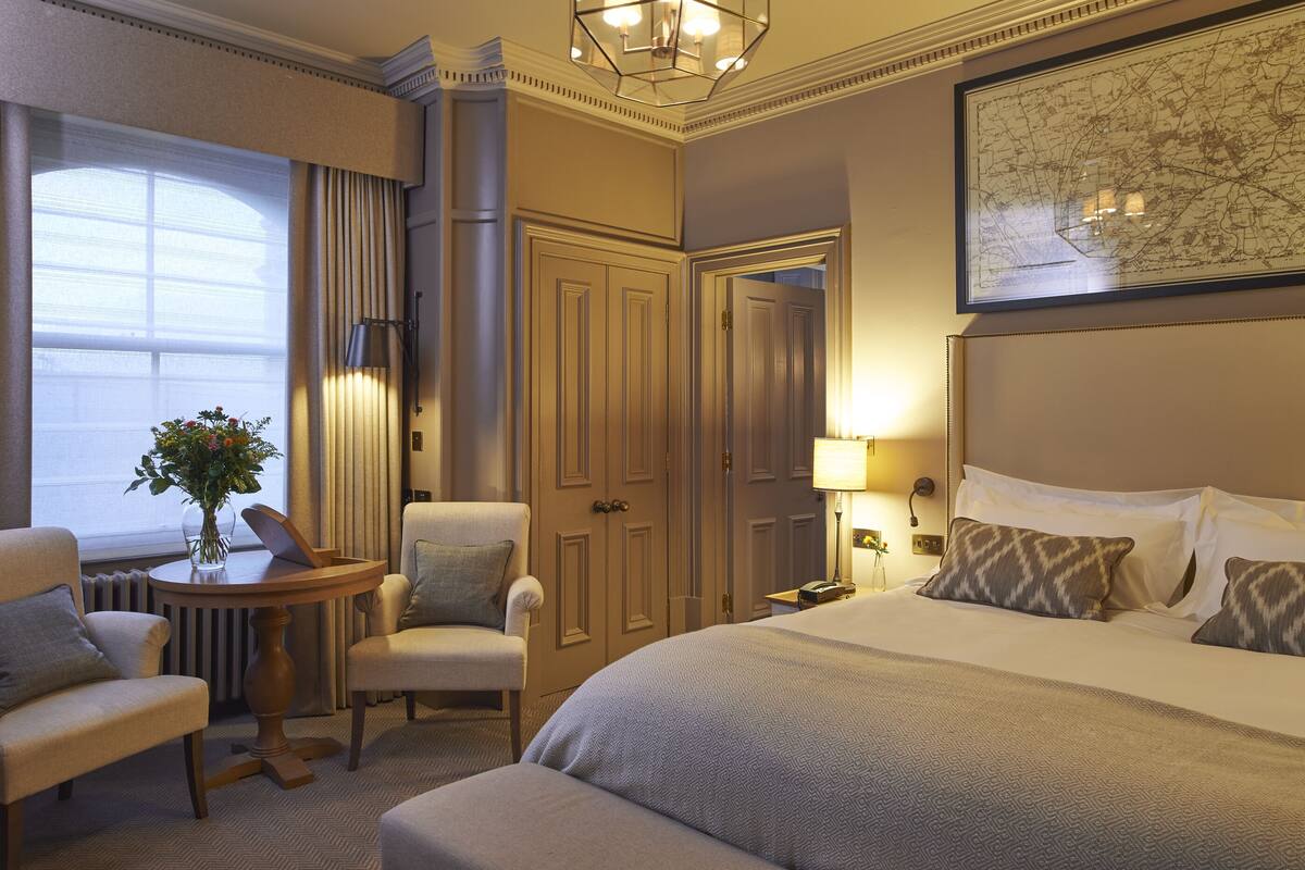 A hotel room with a large bed, two armchairs, a small table with a vase, a lamp, and a map on the wall.