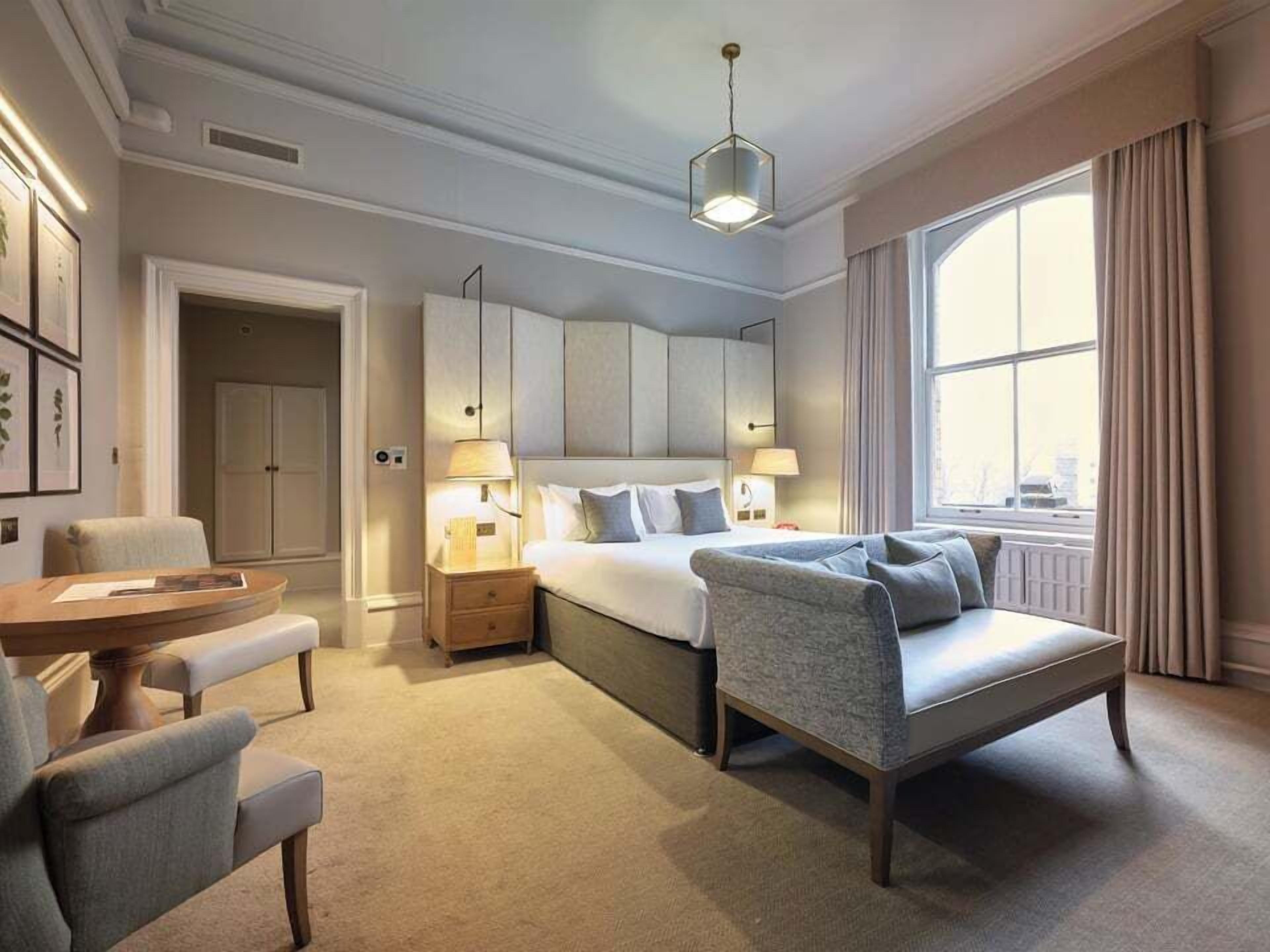 Ein modernes Hotelzimmer mit einem großen Bett, einer Sitzecke mit Tisch und Stühlen, einem Schreibtisch und einem großen Fenster mit Vorhängen.