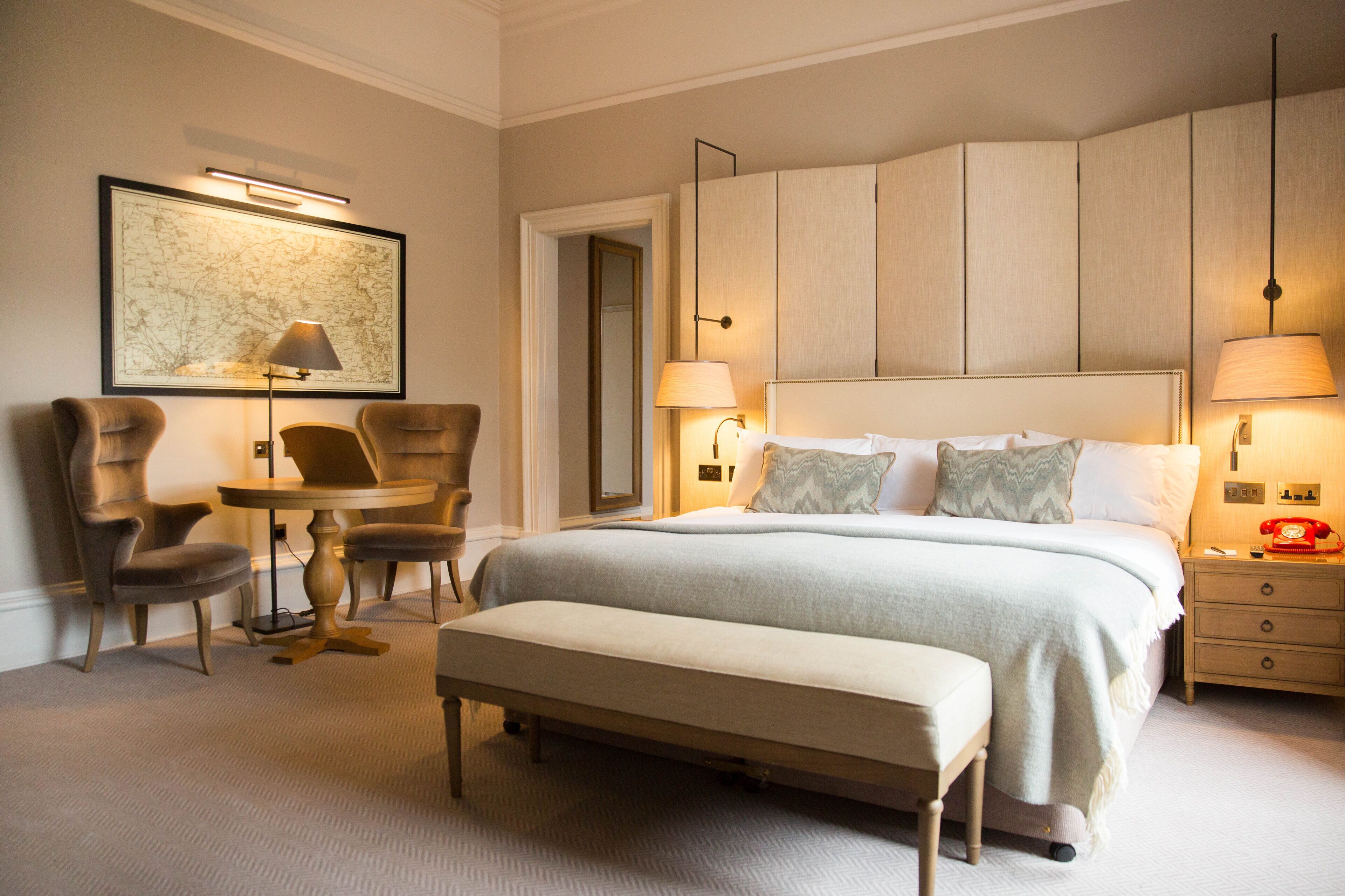 A hotel room with a large bed, two bedside tables with lamps, a bench, a small table with chairs, and a framed artwork on the wall.