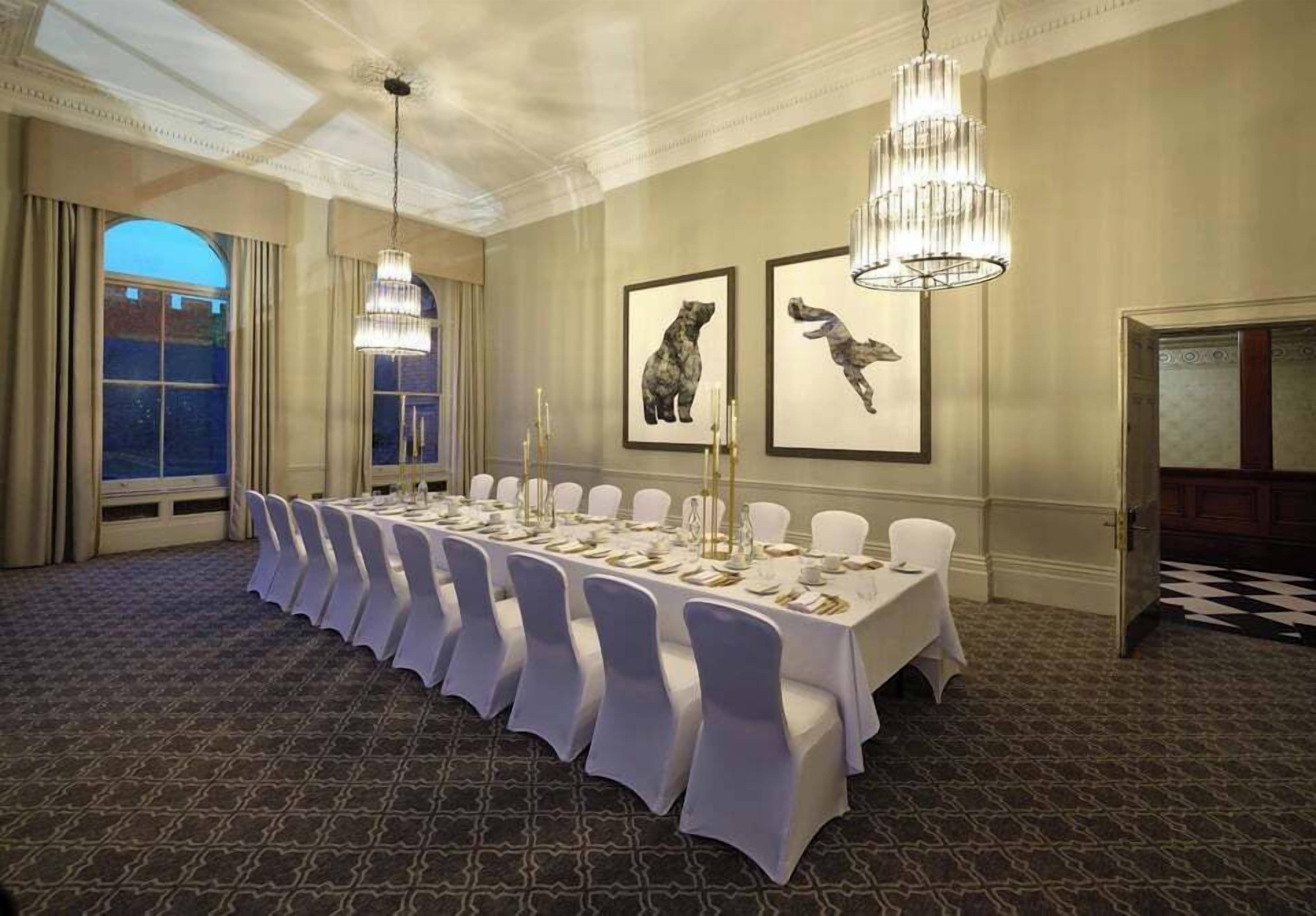 A banquet hall with a long table set for a formal event, chandeliers, framed animal artwork on the walls, and a patterned carpet.