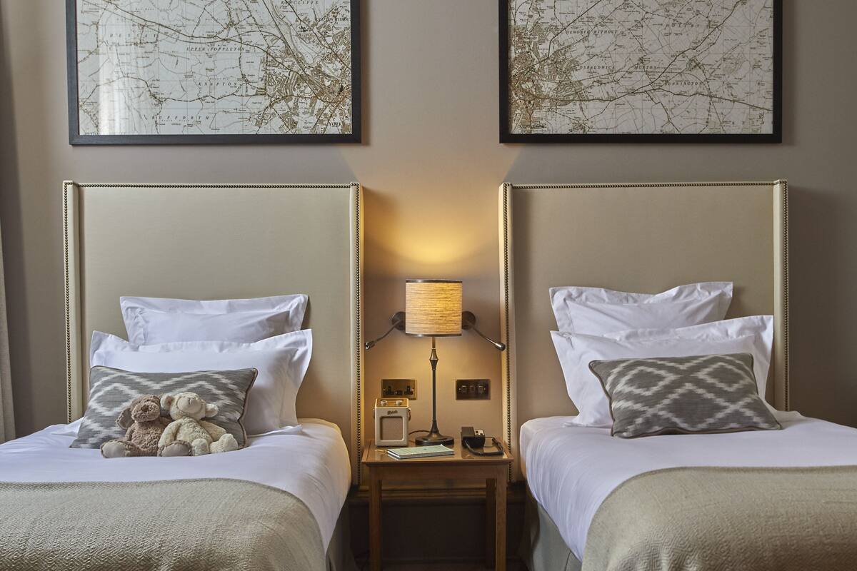 A hotel room with two beds, a nightstand with a lamp, and two framed maps on the wall.