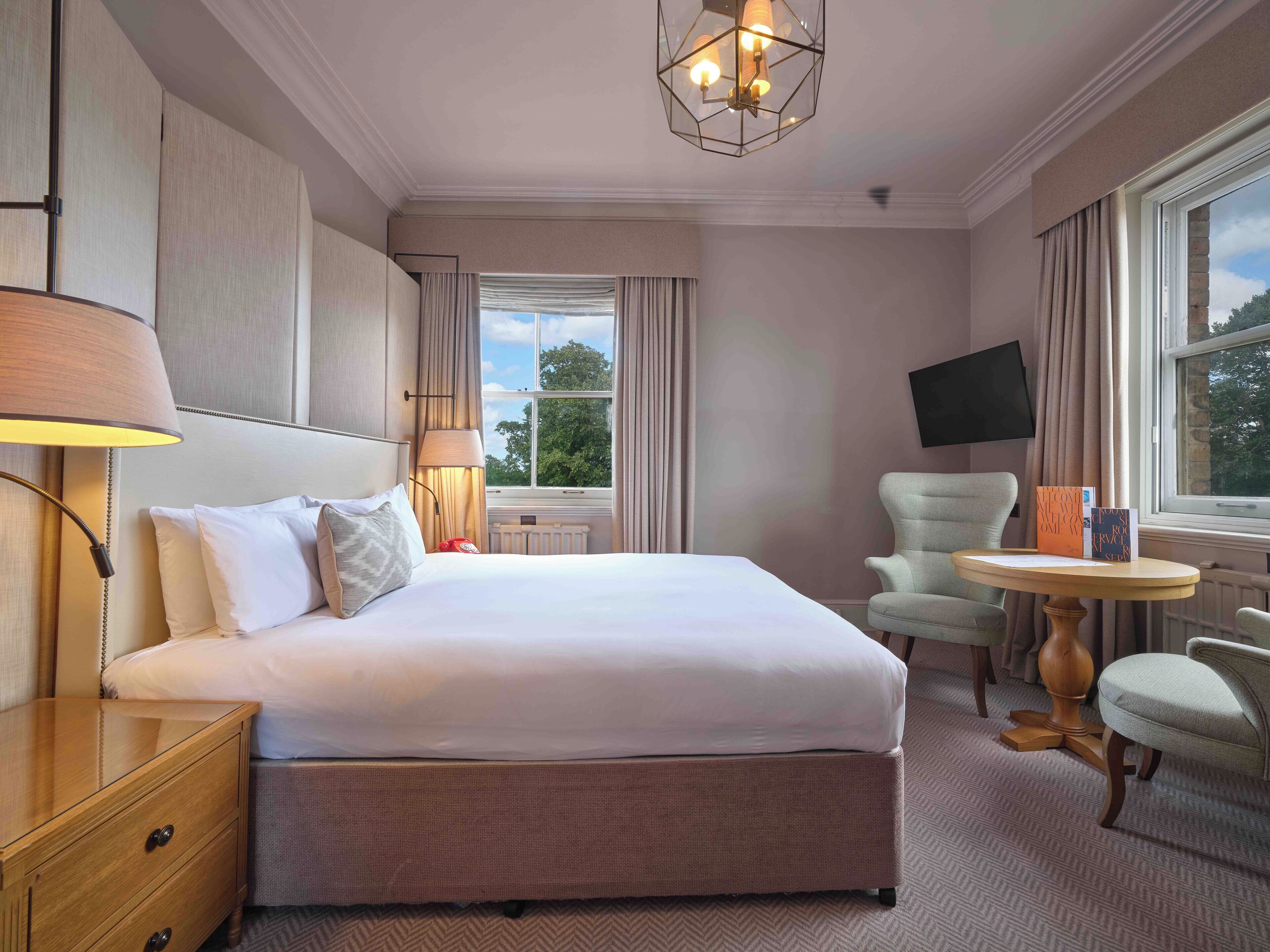 A hotel room with a large bed, a bedside table, a chair, a small table, and a TV mounted on the wall.