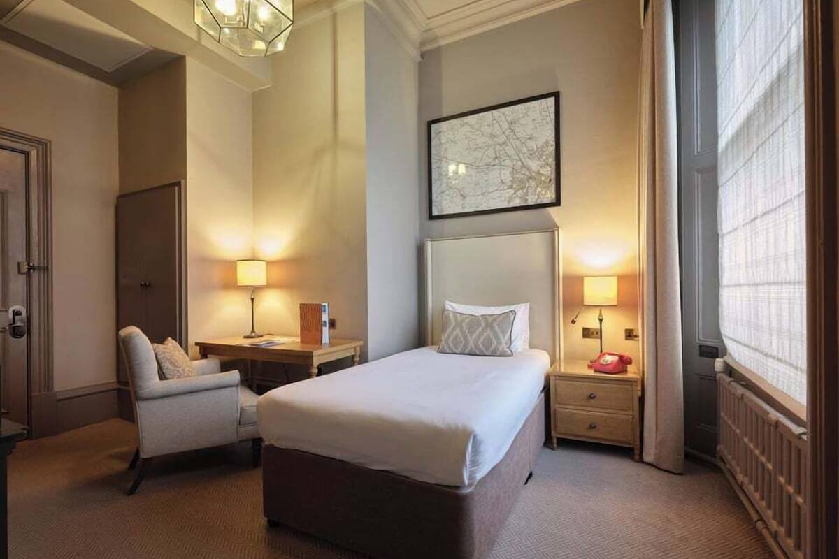 A hotel room with a large bed, a desk, a chair, and a nightstand with a lamp.