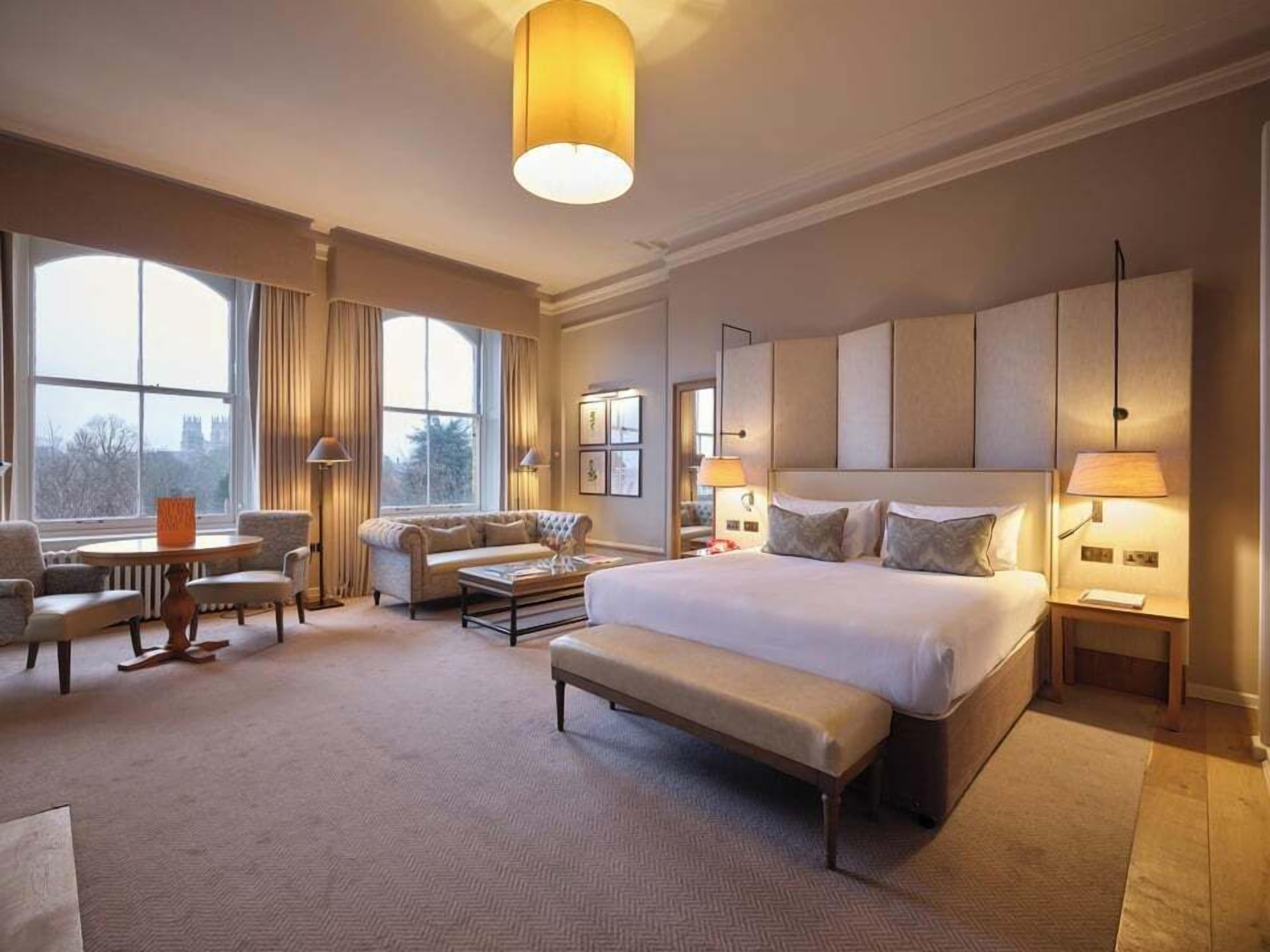 Amplia habitación de hotel con una cama grande, una zona de estar con sofá y sillones, una mesita y un ventanal con vistas al exterior.