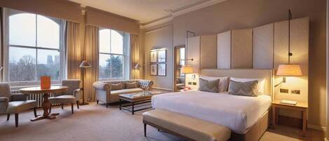 Amplia habitación de hotel con una cama grande, una zona de estar con sofá y sillones, una mesita y un ventanal con vistas al exterior.