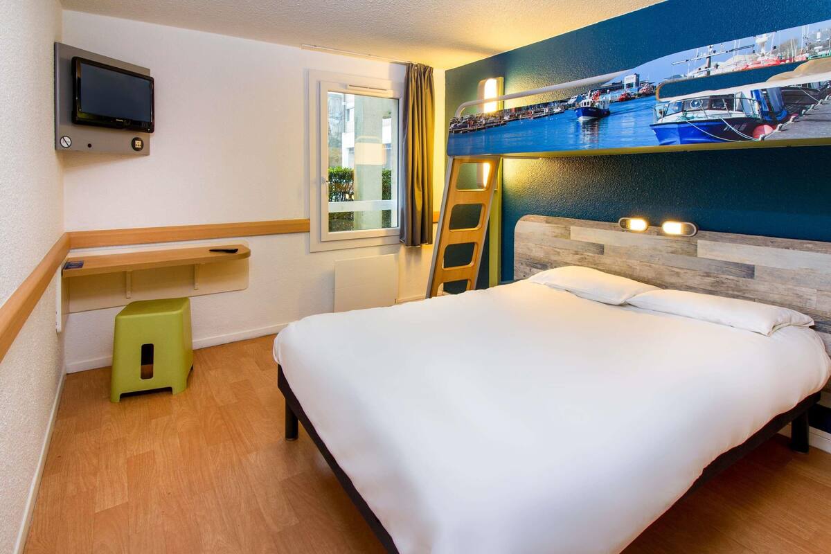 A hotel room with a bed, a TV mounted on the wall, a window, and a wooden floor.