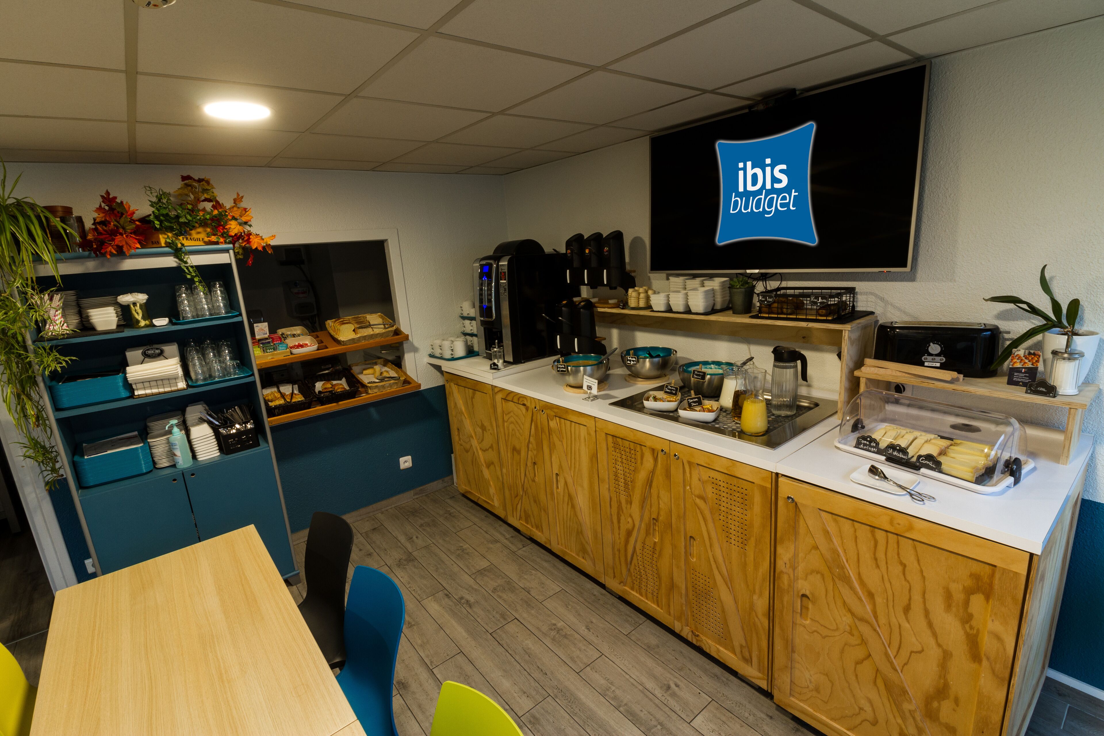 A modern kitchen with a coffee station, a TV displaying 'ibis budget', and a dining area with a table and chairs.