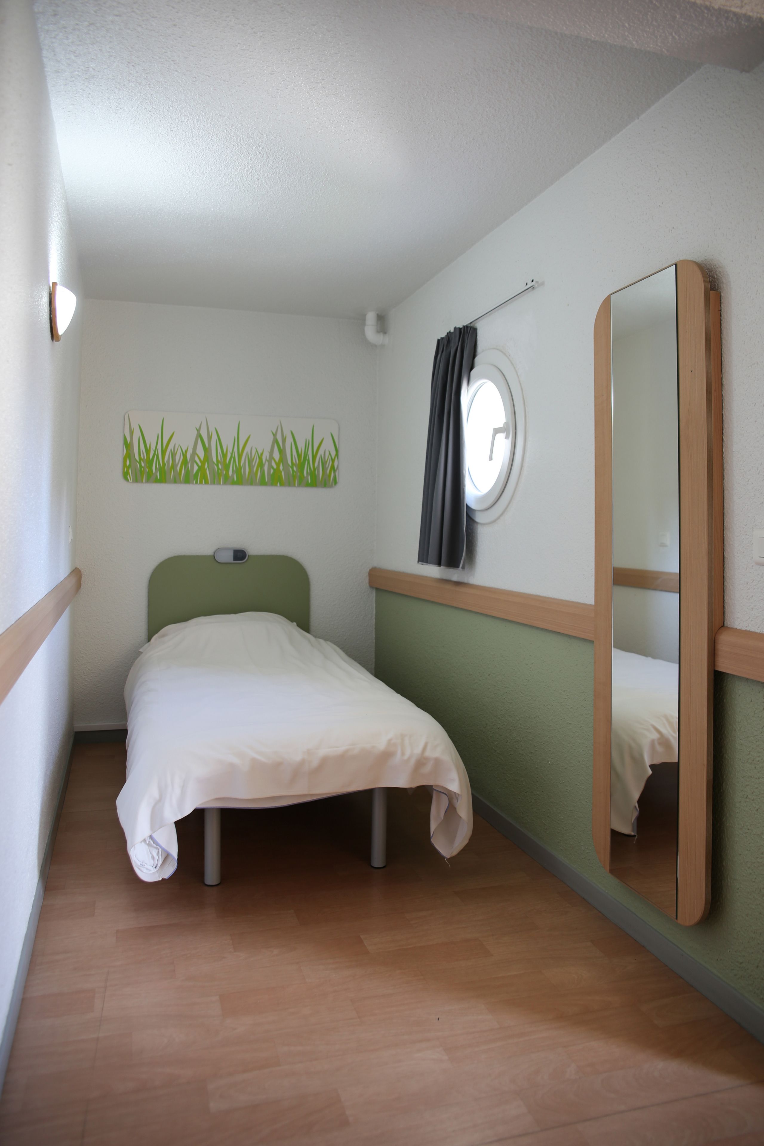 Une petite chambre avec un lit simple, dotée d’une tête de lit verte, d’un miroir rond et d’une horloge fixée au mur.