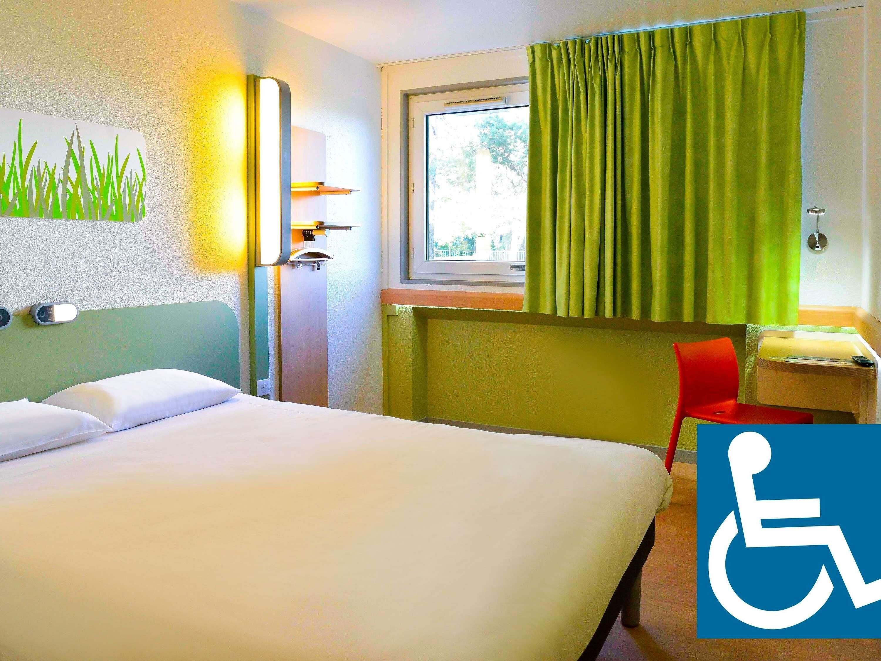 A hotel room with a bed, a desk, a chair, and a window with green curtains. There is a wheelchair accessibility sign on the wall.