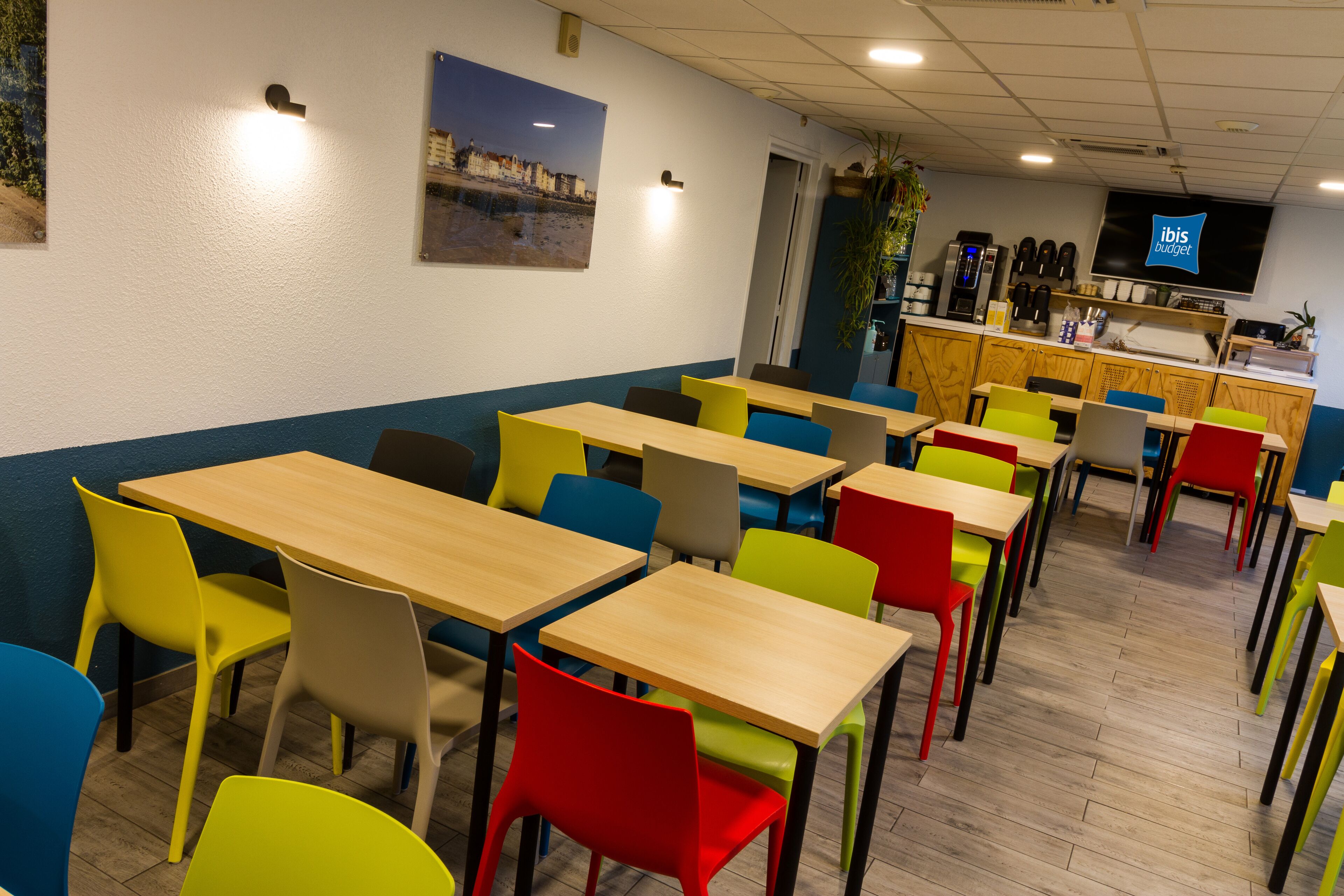 A dining area with multiple tables and chairs, a counter with a coffee machine, and a television screen displaying 'ibis budget'.