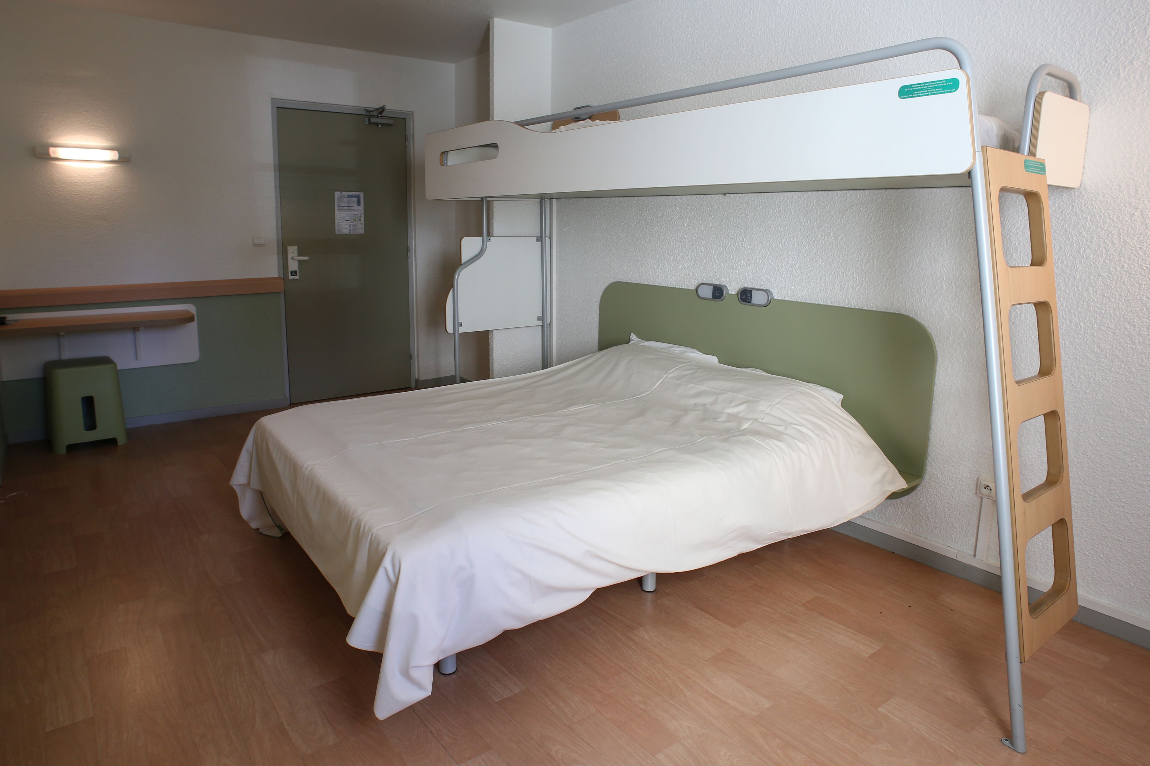 Una cama litera con cabecera verde, escalera de madera y colchón blanco.