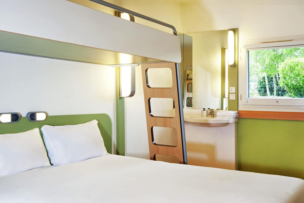 A hotel room with a bed, a bathroom with a sink and mirror, and a window with a view of greenery.