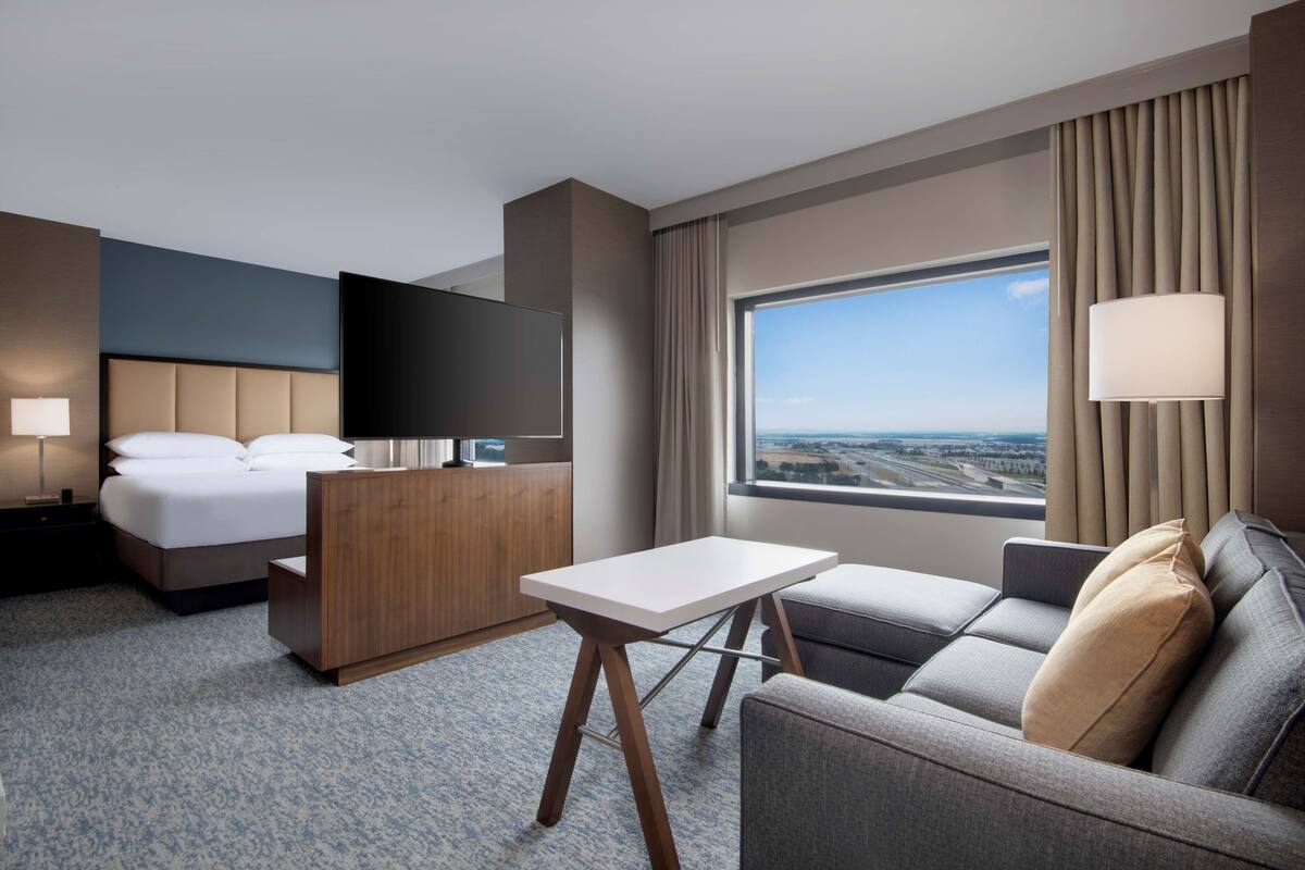 Habitación de hotel con cama, escritorio, sofá, televisión y un amplio ventanal con vistas.