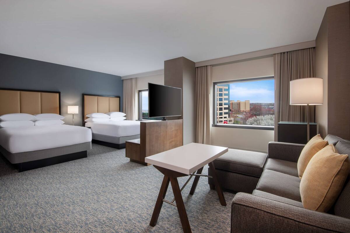 Habitación de hotel con dos camas, un sofá, una mesita, un televisor y una ventana con vistas a la ciudad.