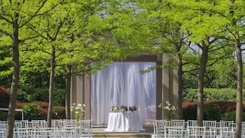 Un lugar para bodas al aire libre con sillas blancas dispuestas en filas, un escenario techado y un telón de fondo de árboles y follaje.