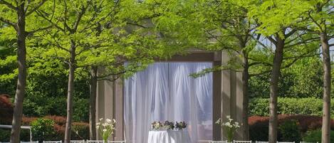 Un lugar para bodas al aire libre con sillas blancas dispuestas en filas, un escenario techado y un telón de fondo de árboles y follaje.