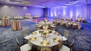 A banquet hall with round tables set for dining, featuring gold and white table settings, and a carpeted floor.