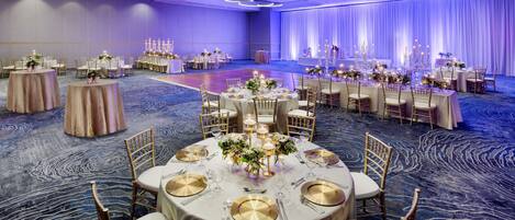 A banquet hall with round tables set for dining, featuring gold and white table settings, and a carpeted floor.