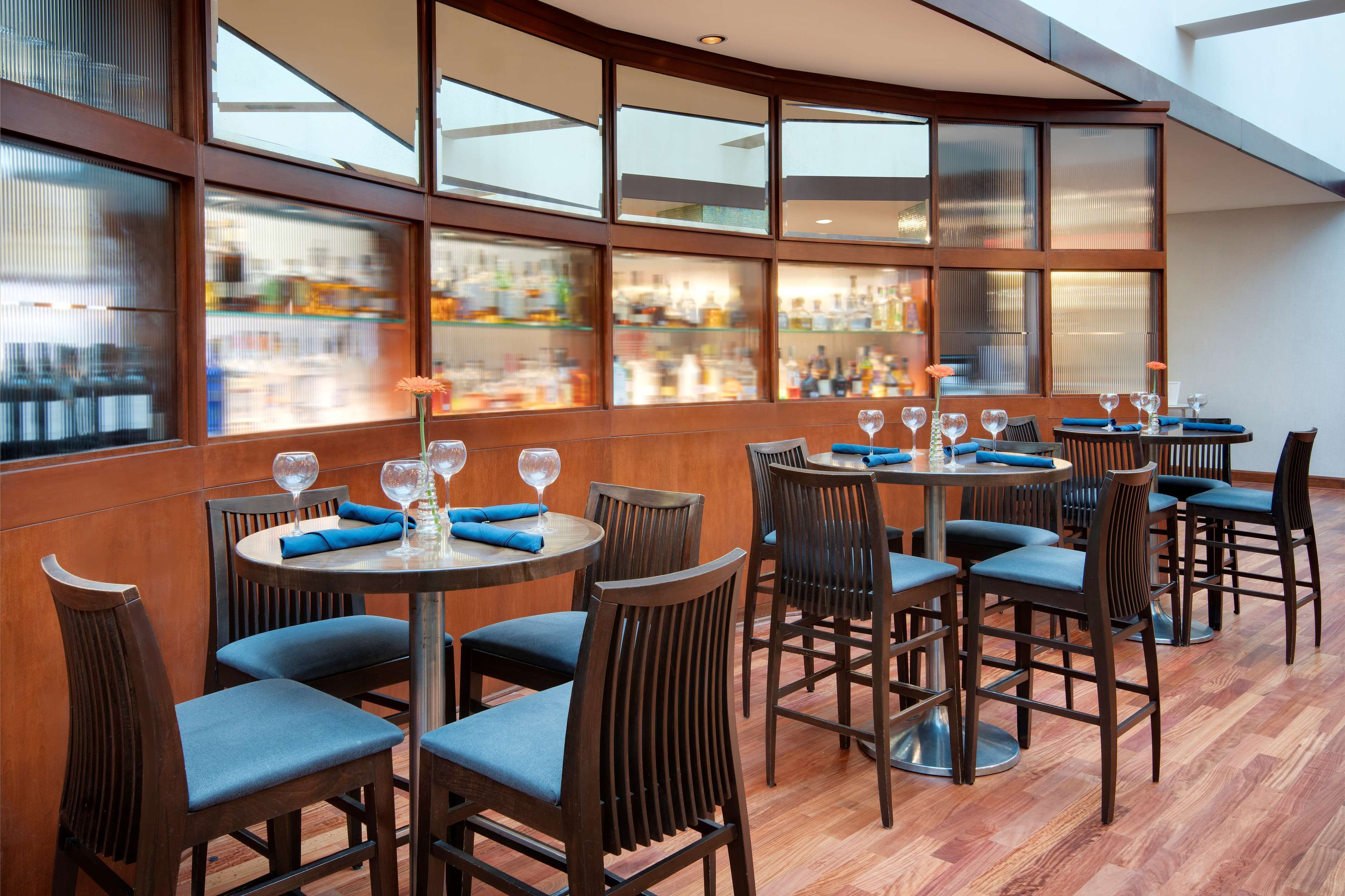A restaurant with round tables, blue cushioned chairs, and a view of a bar through large windows.