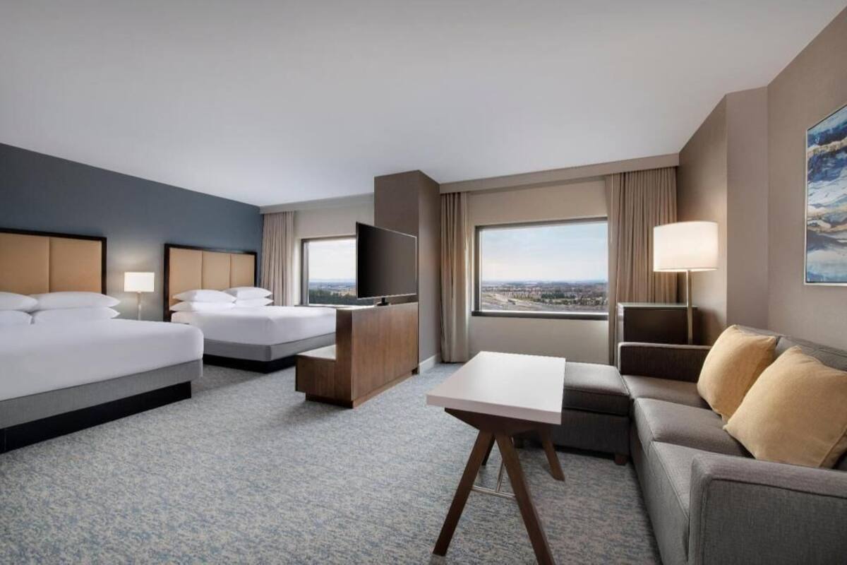 Habitación de hotel con dos camas, televisión, escritorio, sofá y una ventana grande con vista a la ciudad.