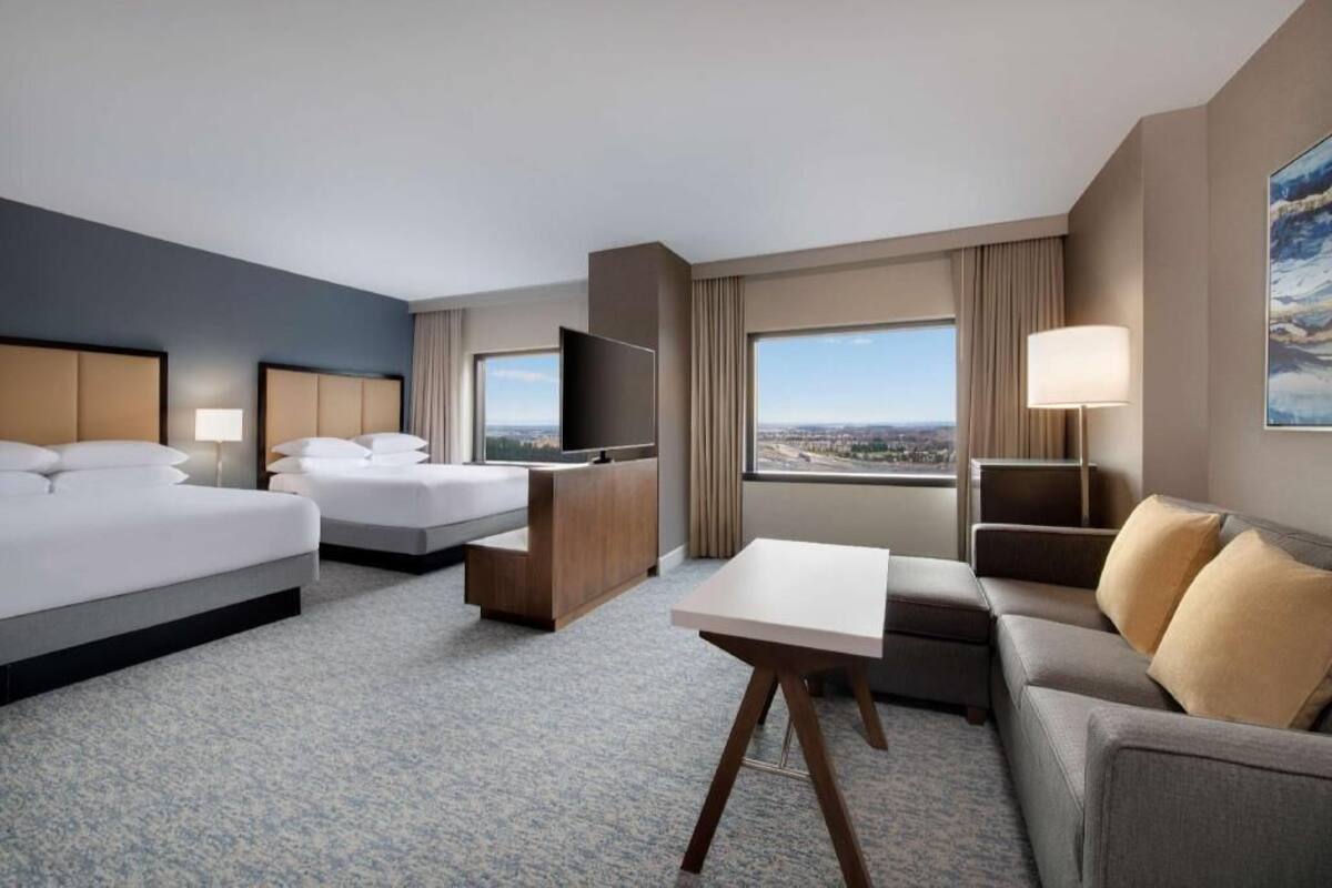 Habitación de hotel con dos camas, un escritorio, un sofá, un televisor y un amplio ventanal con vistas a la ciudad.