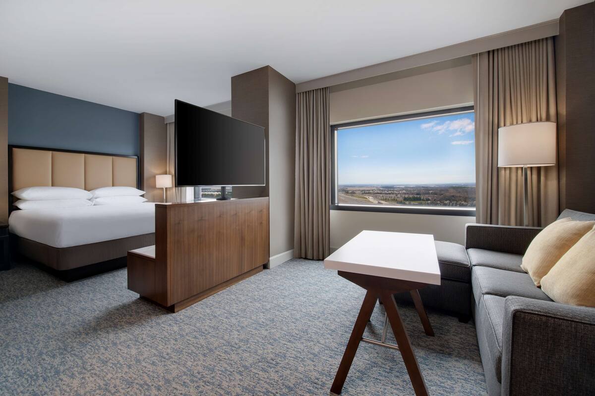 Habitación de hotel con cama, escritorio, sofá, televisor y un amplio ventanal con vista a la ciudad.
