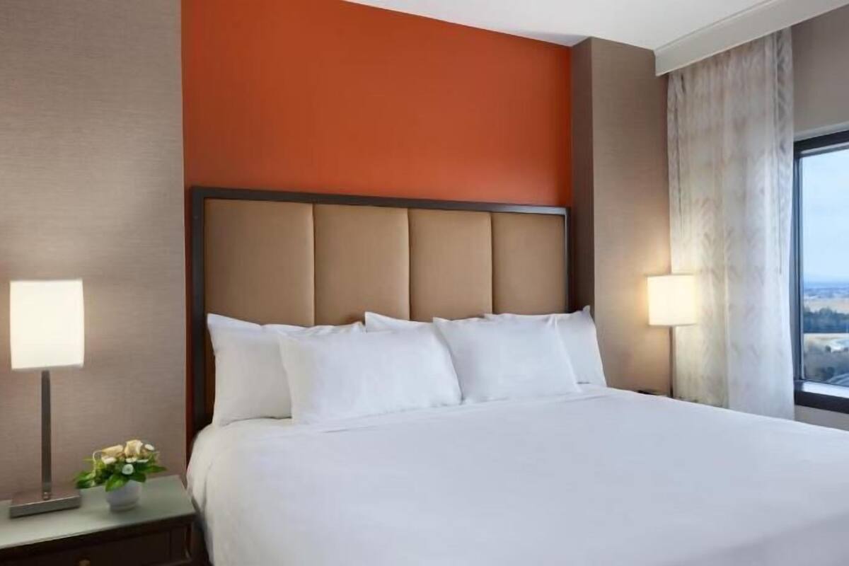 Habitación de hotel con una cama grande, dos lámparas de noche, una mesita de noche con un florero y una ventana con vistas al campo.