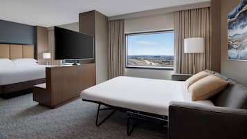 A hotel room with a bed, a sofa, a desk, a TV, and a large window offering a city view.